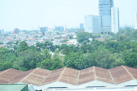 An urban landscape with a blend of natural and architectural elements. In the foreground, several large industrial buildings with brownish roofs are visible, surrounded by lush green trees and vegetation. Behind the greenery, a densely packed residential area with various houses can be seen, leading up to taller modern buildings and skyscrapers in the background.
