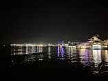 A nighttime waterfront scene with a large yacht illuminated with lights docked to the right. City lights twinkle in the background, reflecting on the calm water.