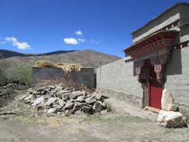 A rustic outdoor setting with a traditional building featuring a decorative doorway. The structure has a flat roof and is made of clay or mud bricks with intricate colorful patterns around the entrance. A pile of rocks is on the ground nearby, and hay is stacked on top of a wall. The landscape is mountainous with a clear blue sky and a few clouds.