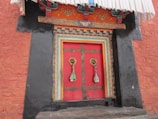 A decorative wooden door is framed by intricate carvings and vibrant colors, featuring a predominant red with golden brass handles, adorned with colorful fabric tassels. The door is set in a textured, reddish-orange wall constructed from stone bricks and highlighted by black painted side panels.