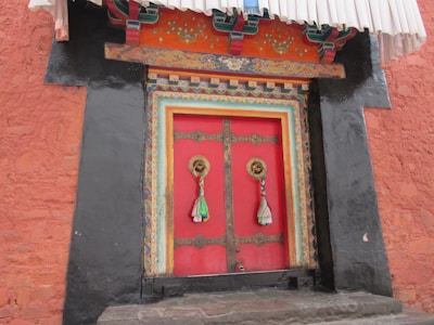 A decorative wooden door is framed by intricate carvings and vibrant colors, featuring a predominant red with golden brass handles, adorned with colorful fabric tassels. The door is set in a textured, reddish-orange wall constructed from stone bricks and highlighted by black painted side panels.