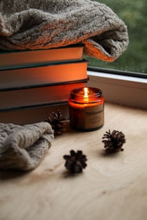 A cozy setup featuring a stack of books with a knitted blanket draped over them. An amber jar candle is lit, emitting a warm glow, surrounded by a few pine cones on a wooden surface. The scene is by the window, adding to the serene and comforting environment.