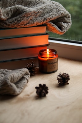 A cozy setup featuring a stack of books with a knitted blanket draped over them. An amber jar candle is lit, emitting a warm glow, surrounded by a few pine cones on a wooden surface. The scene is by the window, adding to the serene and comforting environment.