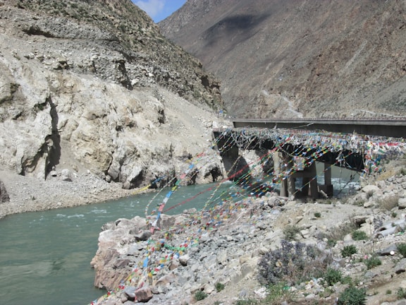 A rugged landscape featuring a bridge draped with colorful prayer flags spanning a river. The rocky cliffs around the river are barren and steep, highlighting the remote and natural environment. The water appears teal and swiftly flowing, contrasting with the brown and gray tones of the surrounding rocks.