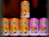 Seven cans of a beverage are arranged, with colorful designs and text in a foreign script on the labels. The cans in the foreground are primarily in orange and pink shades, featuring illustrations of mango and peach with coconuts, suggesting tropical flavors.