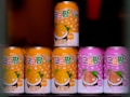 Seven cans of a beverage are arranged, with colorful designs and text in a foreign script on the labels. The cans in the foreground are primarily in orange and pink shades, featuring illustrations of mango and peach with coconuts, suggesting tropical flavors.