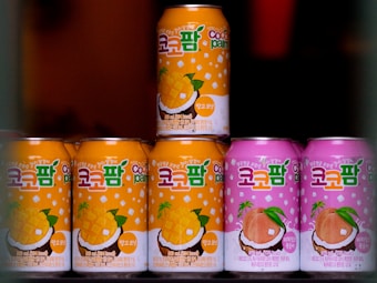 Seven cans of a beverage are arranged, with colorful designs and text in a foreign script on the labels. The cans in the foreground are primarily in orange and pink shades, featuring illustrations of mango and peach with coconuts, suggesting tropical flavors.
