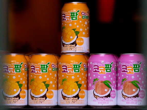 Seven cans of a beverage are arranged, with colorful designs and text in a foreign script on the labels. The cans in the foreground are primarily in orange and pink shades, featuring illustrations of mango and peach with coconuts, suggesting tropical flavors.