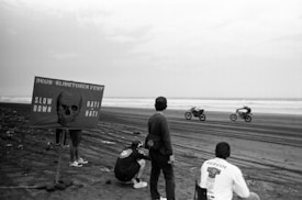 A black and white photo depicts a motorcycle race on a beach. Four individuals are in the foreground, with one sitting on the ground, and another holding a surfboard. A sign with a skull and the text 'DEUS SLIDETOBER FEST SLOW DOWN HATI-HATI' is prominently displayed. Two motorcycles are racing in the background near the shoreline.