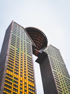 Two tall skyscrapers are connected by a unique circular structure at the top, with a grid of windows in yellow and green shades, extending high into a pale, overcast sky.
