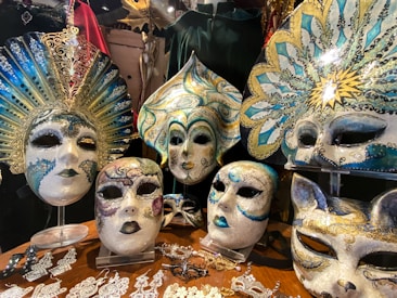 A collection of ornate and colorful Venetian masks is displayed. Each mask features elaborate designs with metallic accents, intricate patterns, and vibrant hues. Some masks have sunburst motifs and others resemble animals, accented with gold and teal. They are placed on a wooden surface alongside delicate lace and beadwork accessories.