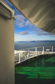 A section of a ship or ferry is visible with a green deck and white railings. The upper part includes part of the ship's structure, casting shadows on the deck. In the background, a calm sea stretches towards distant landmasses under a bright blue sky with scattered clouds.