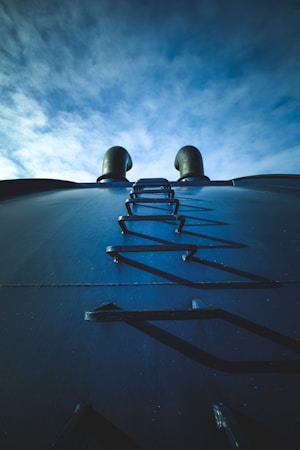 A steel structure with a narrow ladder leading upwards is positioned against a clear blue sky. Two cylindrical exhaust pipes can be seen emerging from the top of the structure, creating an industrial and mechanical impression.