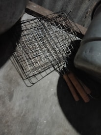 A metal grilling basket with wooden handles is placed on a rough concrete surface, with part of a large pot or container visible. The grid-like structure of the basket casts a shadow on the ground, and the scene is dimly lit, suggesting an outdoor or rustic kitchen setting.