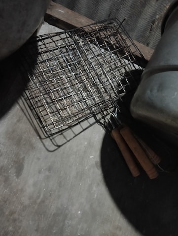 A metal grilling basket with wooden handles is placed on a rough concrete surface, with part of a large pot or container visible. The grid-like structure of the basket casts a shadow on the ground, and the scene is dimly lit, suggesting an outdoor or rustic kitchen setting.