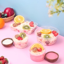 An assortment of fruit salads in clear plastic bowls, topped with slices of orange, kiwi, strawberries, and grapes, arranged on a pink surface. A small wooden bowl with white yogurt and another with pink sauce accompany the salads. A basket of fruits including apples, grapes, and oranges is visible, along with fresh strawberries.