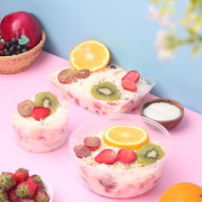 The image features various containers filled with fruit salad, topped with fresh slices of kiwi, orange, strawberries, and grapes. The background is a blend of pink and blue, with additional fruits such as an apple, grapes, and strawberries placed nearby. A small wooden bowl containing a white substance, possibly yogurt or cream, is also present.