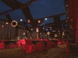 An ornate indoor setting with rich drapery and elegant lighting fixtures. The room features tables and chairs covered in red and black cloths, with decorative lights hanging from above. The ambiance is luxurious with a combination of warm and dark tones.