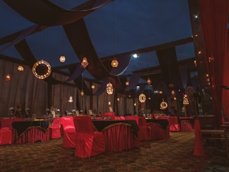 An ornate indoor setting with rich drapery and elegant lighting fixtures. The room features tables and chairs covered in red and black cloths, with decorative lights hanging from above. The ambiance is luxurious with a combination of warm and dark tones.