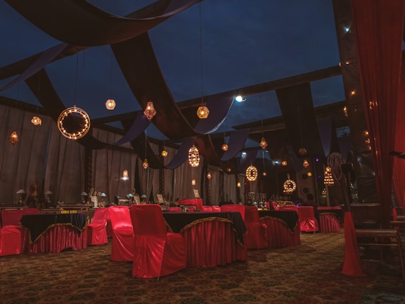 An ornate indoor setting with rich drapery and elegant lighting fixtures. The room features tables and chairs covered in red and black cloths, with decorative lights hanging from above. The ambiance is luxurious with a combination of warm and dark tones.