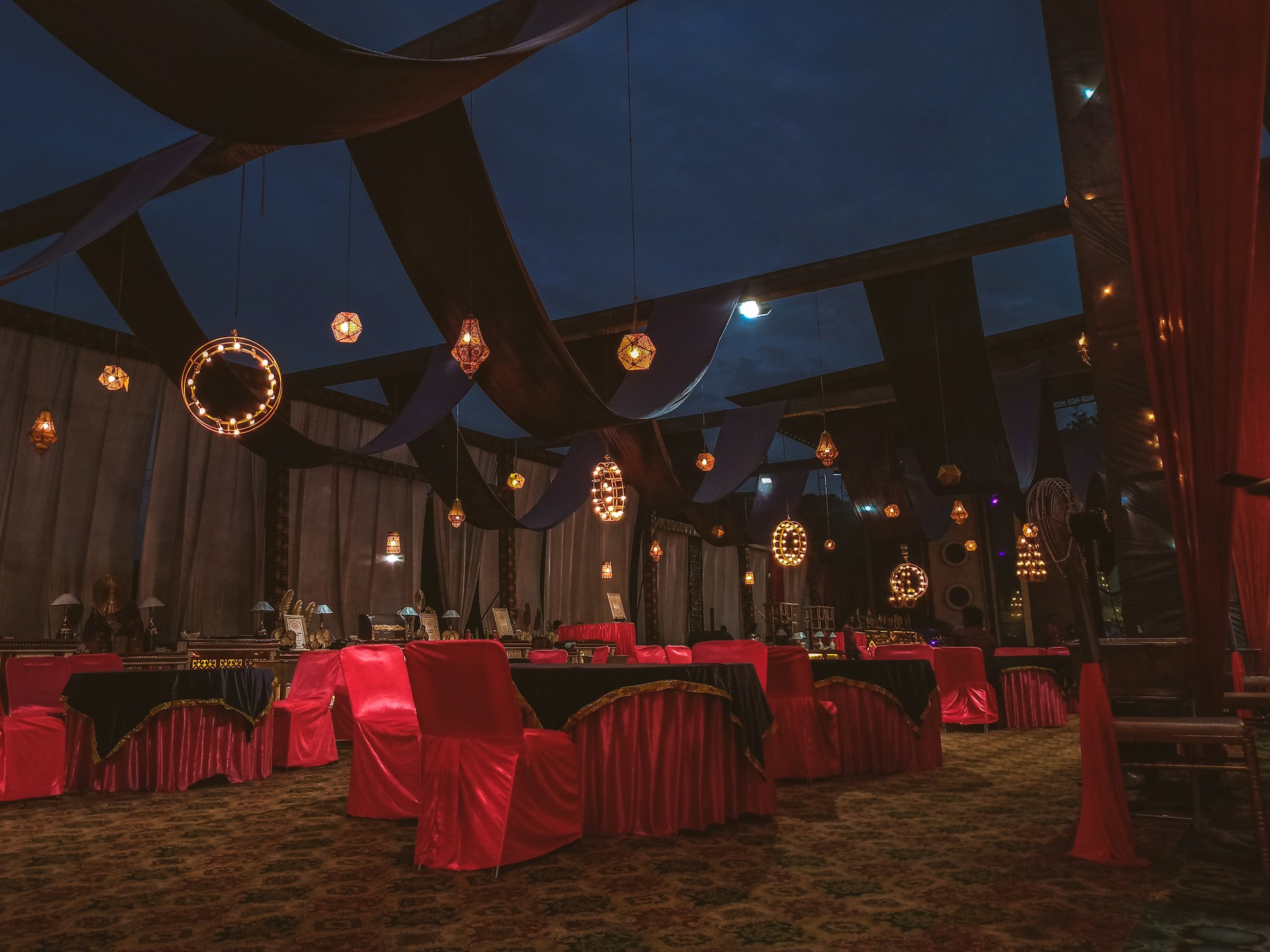 An ornate indoor setting with rich drapery and elegant lighting fixtures. The room features tables and chairs covered in red and black cloths, with decorative lights hanging from above. The ambiance is luxurious with a combination of warm and dark tones.