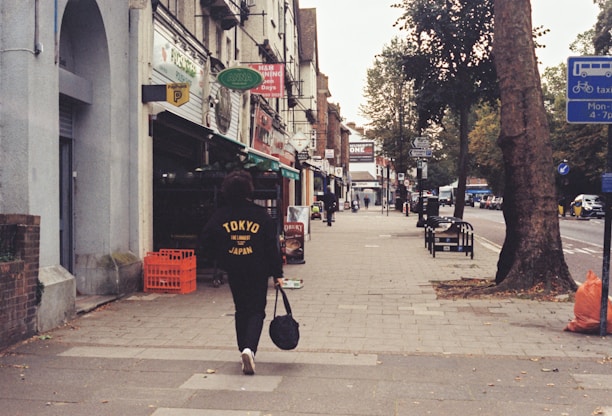 A person wearing a black jacket with 'Tokyo' written on it walks down an urban street lined with trees and shops. There are signs, benches, and a tall tree on the right. The sidewalk appears clean, and there's a red crate near one of the shop entrances.