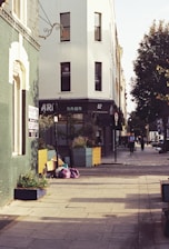 A city street corner featuring a building with white walls and several windows. There are colorful planter boxes with greenery near the building. Some bags and cardboard boxes are placed on the sidewalk. A green building is adjacent to it with a to-let sign. The street is lined with trees and a few pedestrians are walking along the sidewalk in the distance.