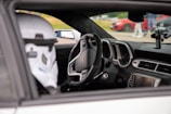 Inside of a Chevrolet car with focus on the driver's seat. The steering wheel features the Chevrolet logo, and there is a white helmet resembling a Stormtrooper helmet from Star Wars on the seat. The dashboard and interior are clearly visible including the speedometer and a mounted device. Outside, a red car and people are visible in a blurred background.
