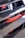 A partial view of a car engine with visible red engine components partially obscured by metallic slats. The focus is on the vibrant red parts with some text visible, indicating branding or specifications, and there are various wires and attachments around it.
