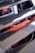 A partial view of a car engine with visible red engine components partially obscured by metallic slats. The focus is on the vibrant red parts with some text visible, indicating branding or specifications, and there are various wires and attachments around it.