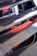 A partial view of a car engine with visible red engine components partially obscured by metallic slats. The focus is on the vibrant red parts with some text visible, indicating branding or specifications, and there are various wires and attachments around it.