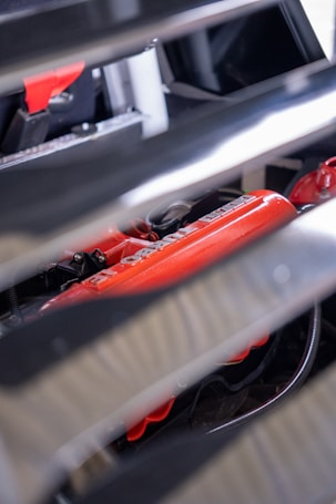 A partial view of a car engine with visible red engine components partially obscured by metallic slats. The focus is on the vibrant red parts with some text visible, indicating branding or specifications, and there are various wires and attachments around it.