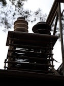A close-up view of an industrial structure with metal rods and two cylindrical objects, possibly insulators, on top. The background includes branches and leaves of a tree silhouetted against a bright sky.