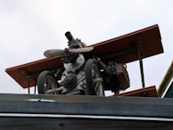 A mechanical structure resembling a small aircraft or drone, mounted on a metallic platform. It includes components such as a propeller, wheels, and what appears to be parts of an engine or machinery. The structure has a worn, industrial look with some rust visible on the metal parts.