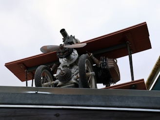 A mechanical structure resembling a small aircraft or drone, mounted on a metallic platform. It includes components such as a propeller, wheels, and what appears to be parts of an engine or machinery. The structure has a worn, industrial look with some rust visible on the metal parts.