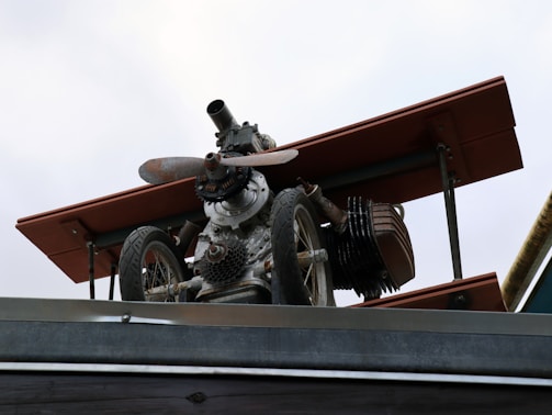A mechanical structure resembling a small aircraft or drone, mounted on a metallic platform. It includes components such as a propeller, wheels, and what appears to be parts of an engine or machinery. The structure has a worn, industrial look with some rust visible on the metal parts.