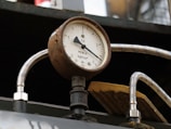 A close-up view of an old, rusty pressure gauge, displaying measurements in kilograms per square centimeter. The gauge is connected to shiny, braided metal hoses and set against a slightly blurred background.