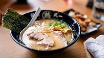 A bowl of ramen with rich broth, slices of pork, green onions, and seaweed. The dish includes mushrooms and other garnishes, with a white spoon resting in the bowl. A side dish and a rolled towel are visible on the wooden table.