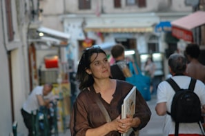 A woman is walking through a busy street holding a book. The background features several other people, storefronts, and a café setting, indicating a lively urban environment. The architecture and signs suggest a European street scene.