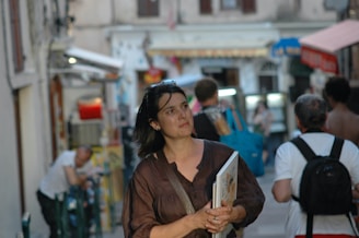 A woman is walking through a busy street holding a book. The background features several other people, storefronts, and a café setting, indicating a lively urban environment. The architecture and signs suggest a European street scene.