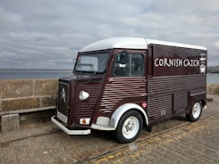 A vintage-style food truck with a dark red and white exterior is parked on a cobblestone pavement by the seaside. The truck has the words 'Cornish Catch' painted on the side, with a small logo of a crab. The background shows a stone wall and a cloudy sky over the ocean.