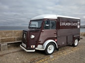 A vintage-style food truck with a dark red and white exterior is parked on a cobblestone pavement by the seaside. The truck has the words 'Cornish Catch' painted on the side, with a small logo of a crab. The background shows a stone wall and a cloudy sky over the ocean.