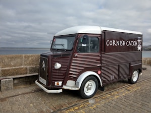 A vintage-style food truck with a dark red and white exterior is parked on a cobblestone pavement by the seaside. The truck has the words 'Cornish Catch' painted on the side, with a small logo of a crab. The background shows a stone wall and a cloudy sky over the ocean.