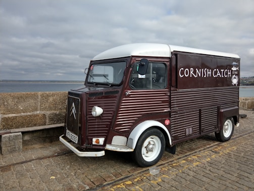 A vintage-style food truck with a dark red and white exterior is parked on a cobblestone pavement by the seaside. The truck has the words 'Cornish Catch' painted on the side, with a small logo of a crab. The background shows a stone wall and a cloudy sky over the ocean.