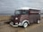 A vintage-style food truck with a dark red and white exterior is parked on a cobblestone pavement by the seaside. The truck has the words 'Cornish Catch' painted on the side, with a small logo of a crab. The background shows a stone wall and a cloudy sky over the ocean.