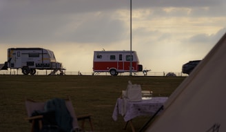 A scenic outdoor setting with two trailers parked on a grassy field during a cloudy day. One trailer is red and white, while the other is grey with yellow accents. A table with a cloth and some items is visible in the foreground, along with a chair. The sky is overcast, casting a serene and calm atmosphere.