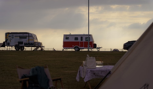 A scenic outdoor setting with two trailers parked on a grassy field during a cloudy day. One trailer is red and white, while the other is grey with yellow accents. A table with a cloth and some items is visible in the foreground, along with a chair. The sky is overcast, casting a serene and calm atmosphere.