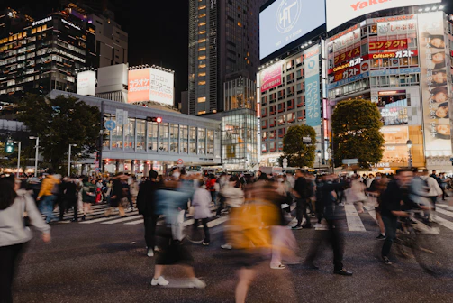 A dynamic 4x6 shot of a bustling city street with people crossing and colorful storefronts.