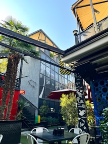 An outdoor setting with modern architecture, featuring a patio area adorned with greenery and palm trees. The structures are mostly glass and metal, displaying geometric shapes. Red accents, including umbrellas and signs, contrast with the natural elements. Visible furniture includes tables and chairs, suggesting a casual, relaxing environment.