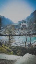 A serene mountain village with houses nestled among bare trees, with a clear river flowing near rocky banks. In the background, forested hills are visible under a bright, hazy sky.